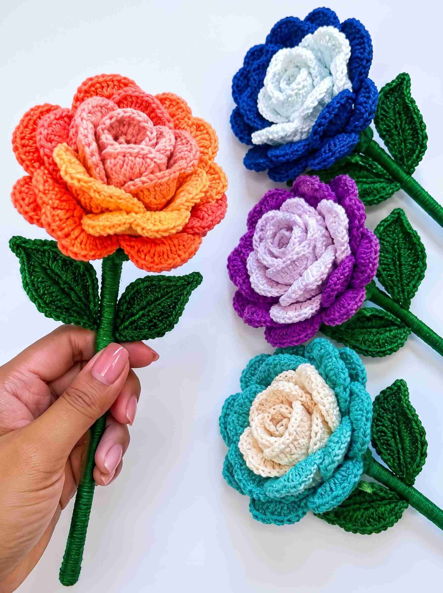 Colorful handmade crochet rose flowers with layered orange, blue, purple, and turquoise petals, green leaves, and wrapped stems.